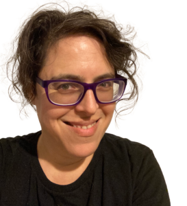an image of phoenix, the poet, a Jewish person with olive skin and wild brown hair and big purple-framed glasses. Xe is smiling a the camera. Xe is wearing a simple black top.